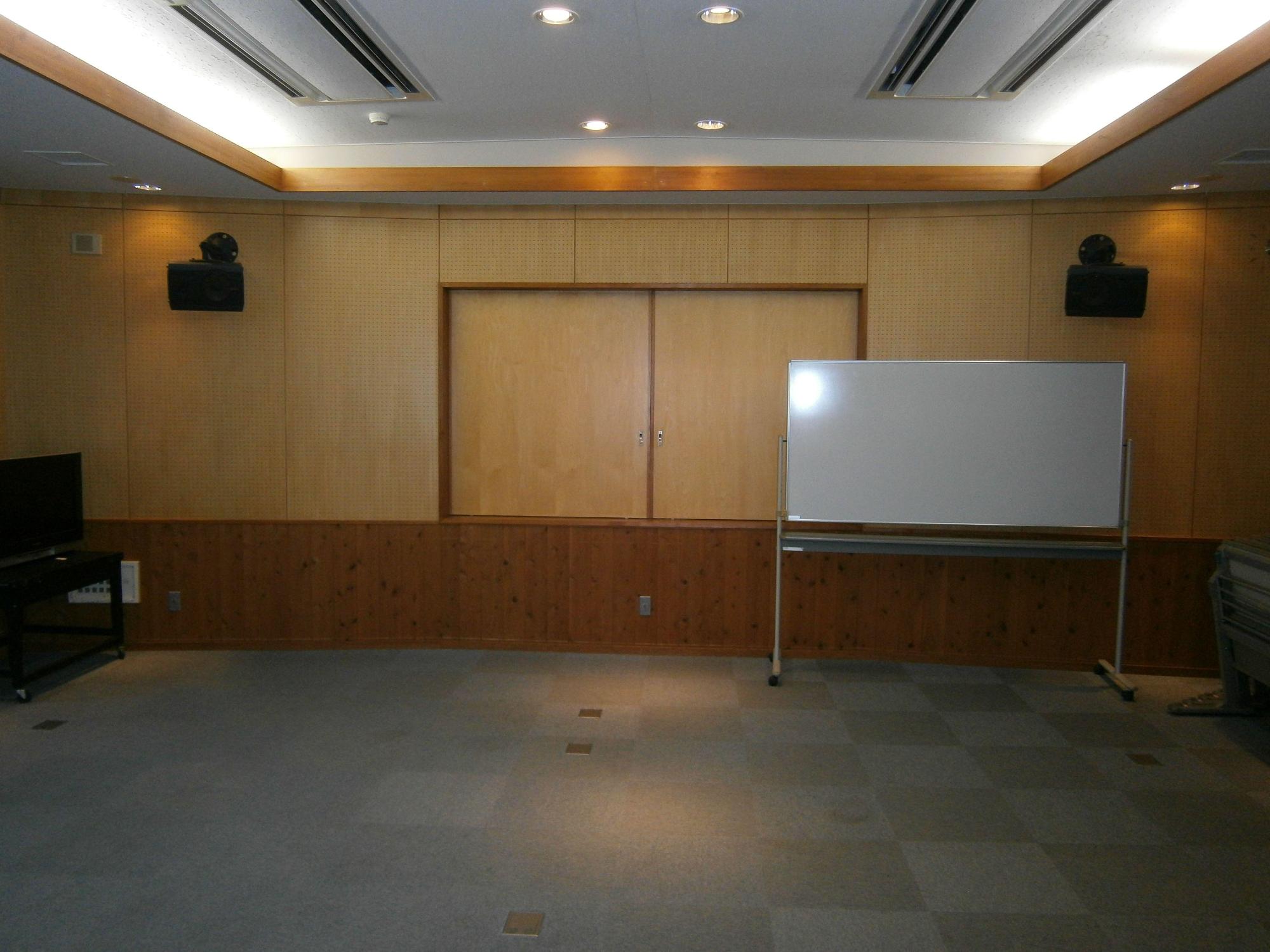 左右の壁上部にスピーカーが設置され、右下にホワイトボード、左に液晶テレビのある視聴覚室の写真