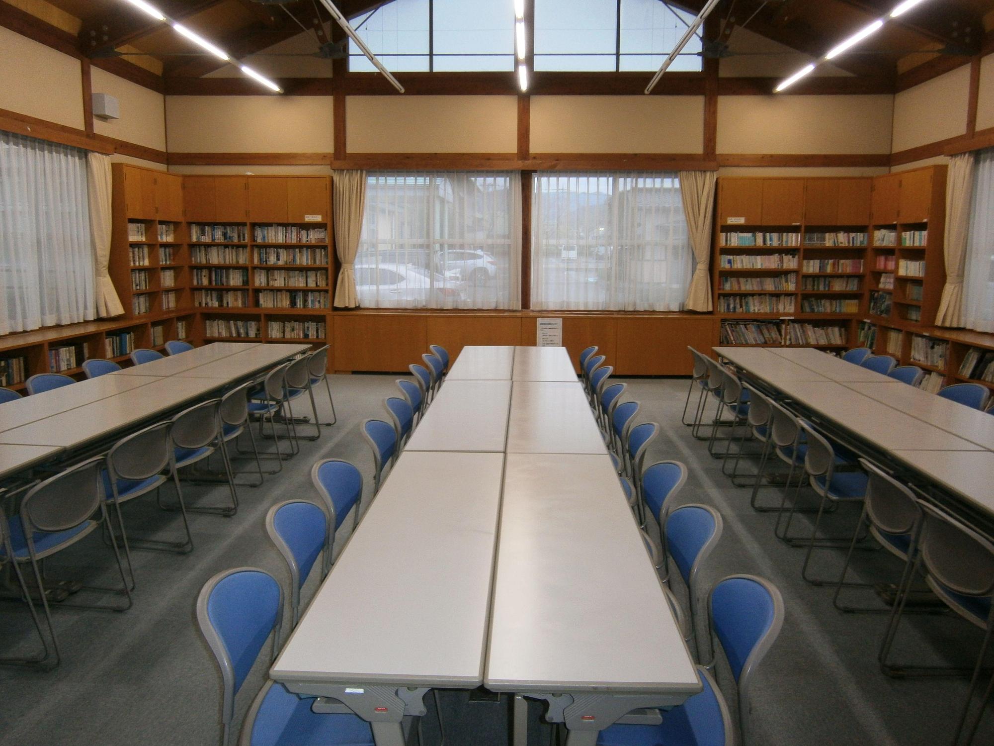 左右と奥にレースカーテンが引いてある腰高窓、窓と窓の間と窓の下に本が並び、3列に向かいあって机と椅子が設置されている図書室の写真
