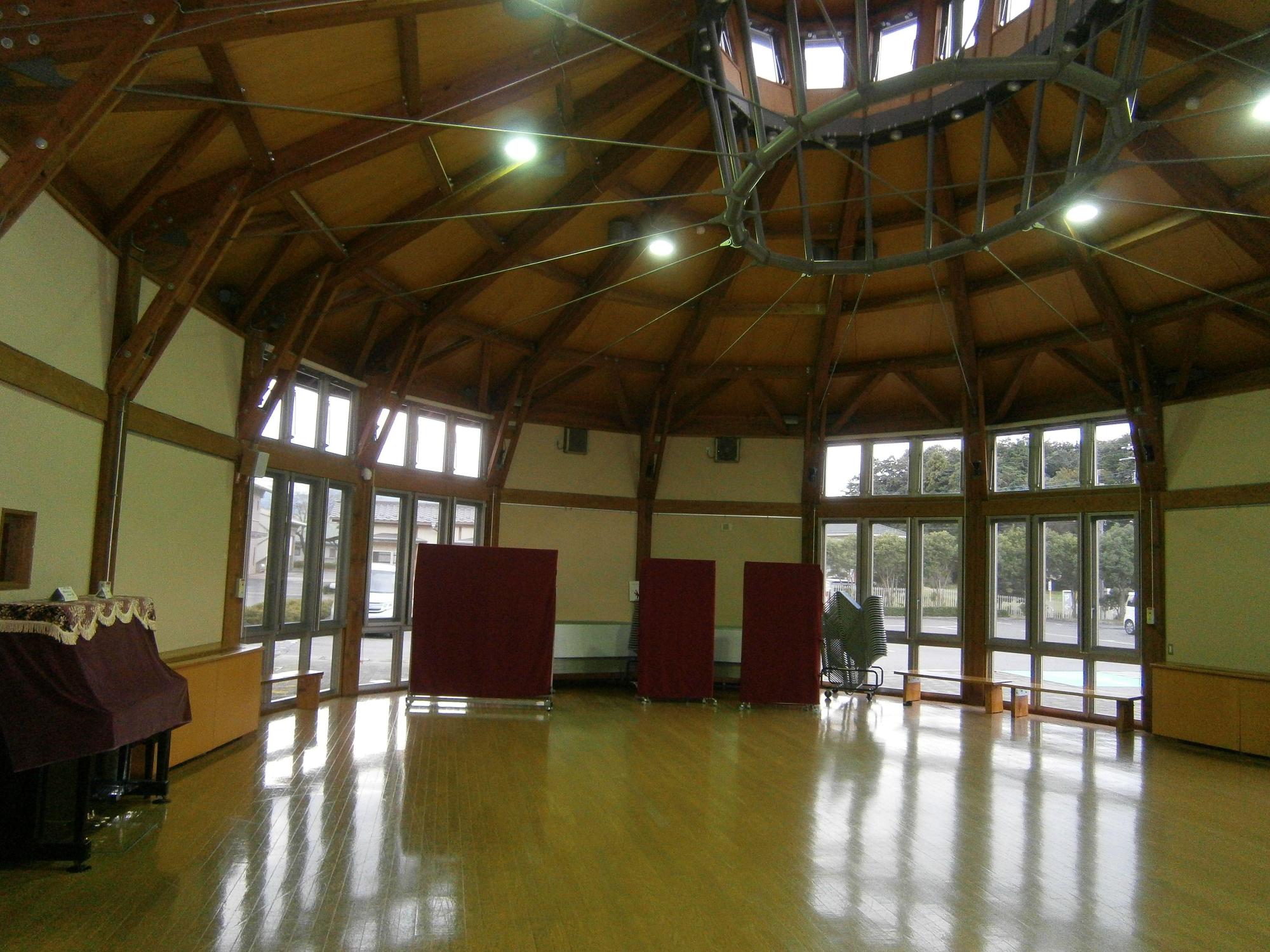 円形の室内左右と天井に窓が施されている明るい室内で、窓の前にベンチ、左側にアップライトピアノが設置されているコミュニティルームゆずの写真