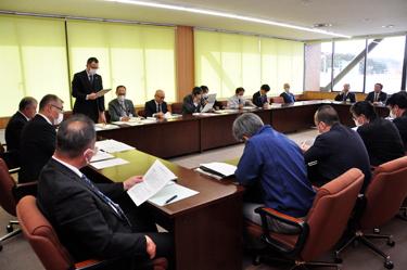 テーブルが口の字型に並べられた会議室で会議が行われており、ブラインド側の席の男性が起立して資料を見ながら話をしている写真