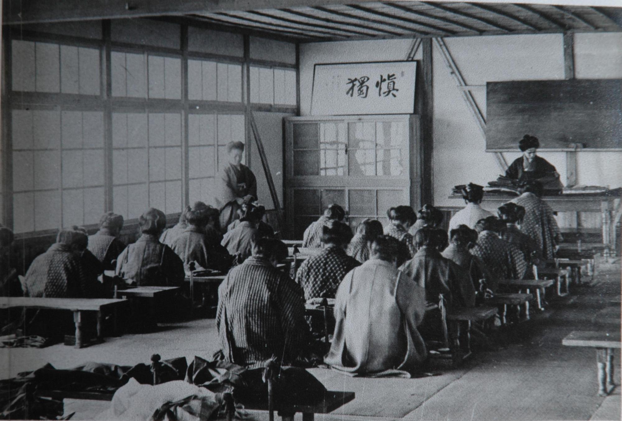 黒板の前や左窓側に教師が立ち、着物を着た生徒が畳の上に並べた長机にそれぞれ正座して座り、授業を受けている白黒写真