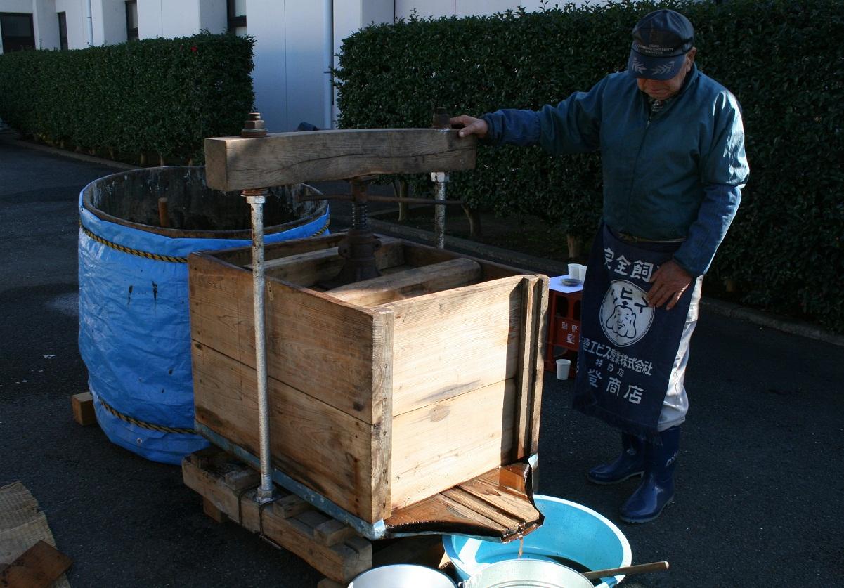 木製の四角形の醤油圧搾機の右側に水色の容器を置いて、醤油を絞り出している現在の写真