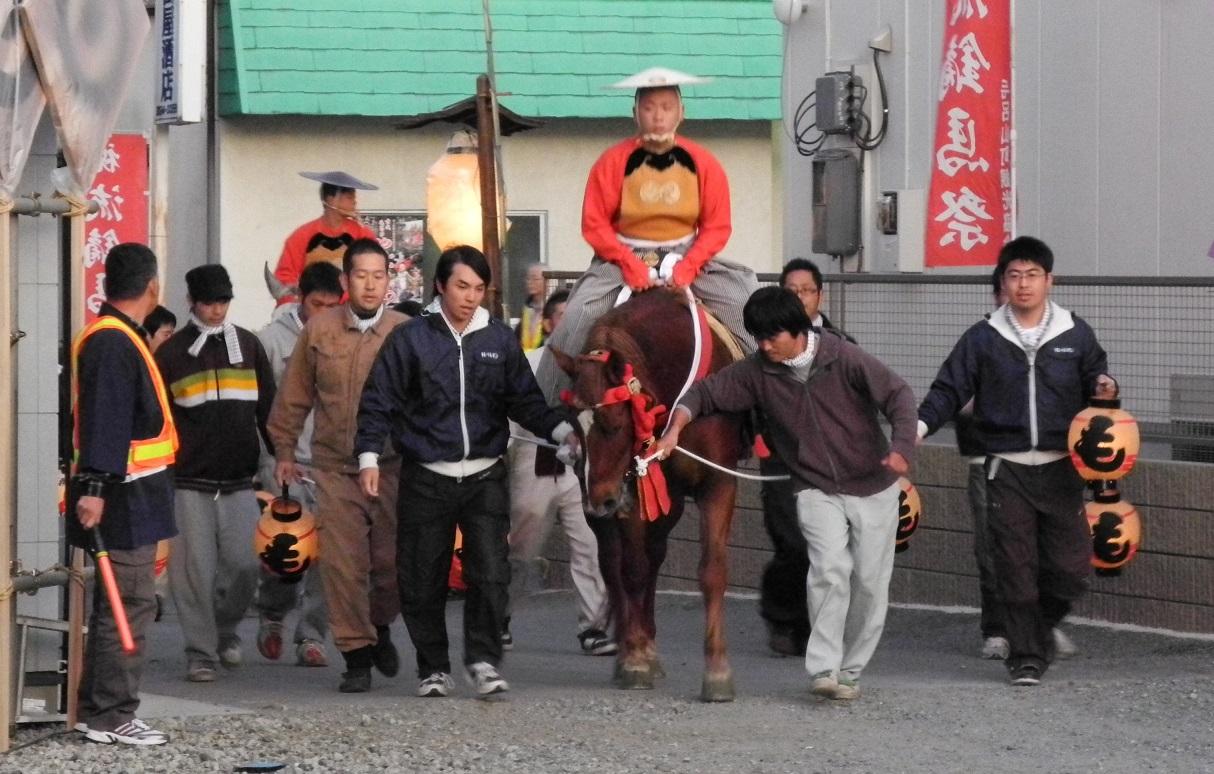 衣装を着て馬に乗った2名の男性の周りを、綱を引っ張っる男性や提灯を持って歩いている人達の写真