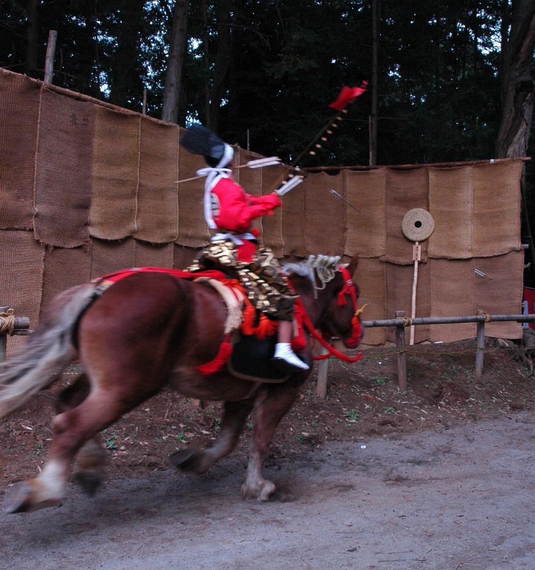 衣装を身にまとった男性が、走っている馬に乗りながら的板に向かって矢を射ている流鏑馬の写真