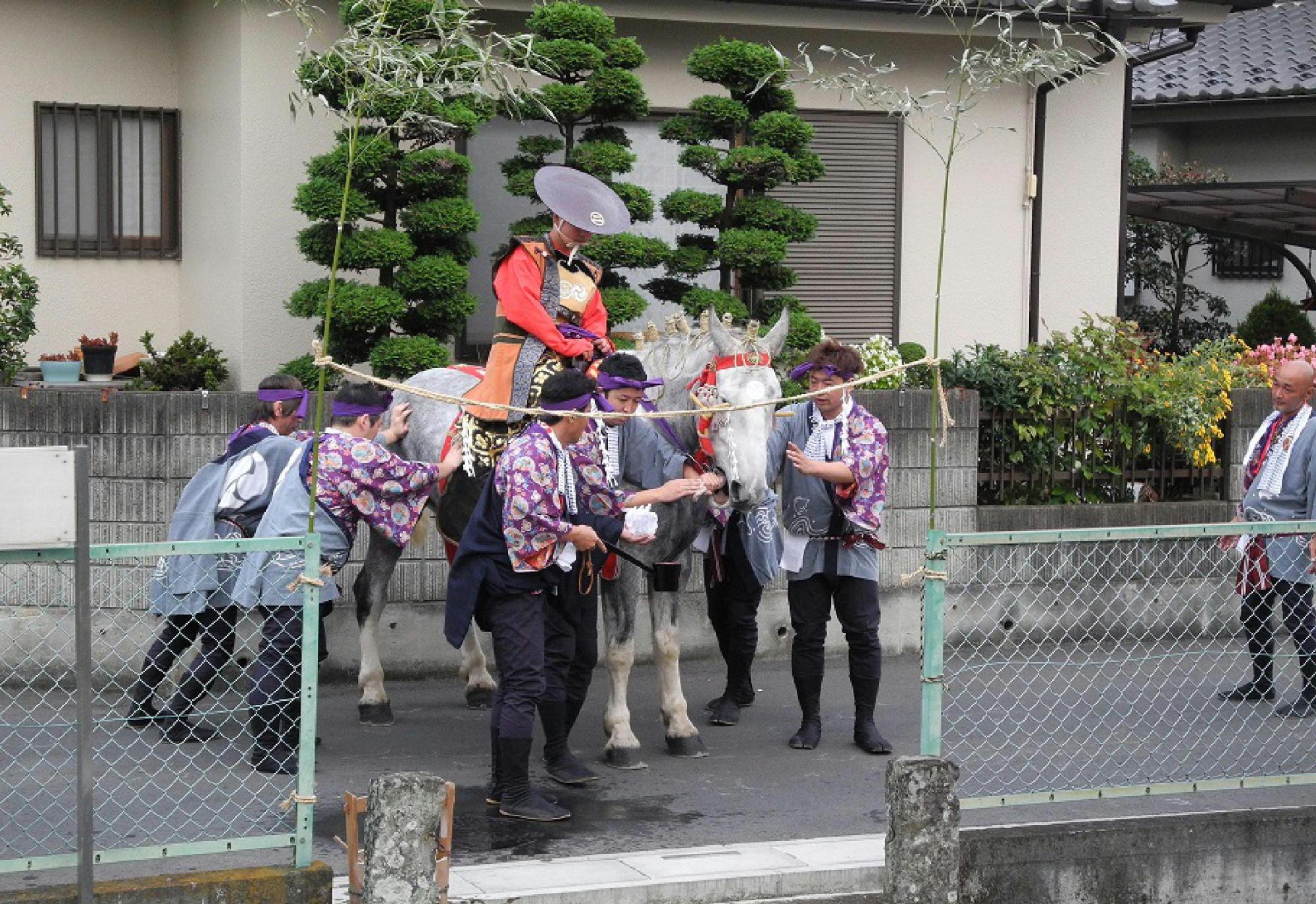 入口の柵に2つの笹を差し綱で繋いだ前に馬を止めて、両側の男性が馬の口をすすいでいる写真