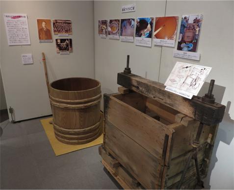 壁に醤油づくりの工程の写真や昔の職人の写真が掲示され、手前に大きな樽などが展示されている写真