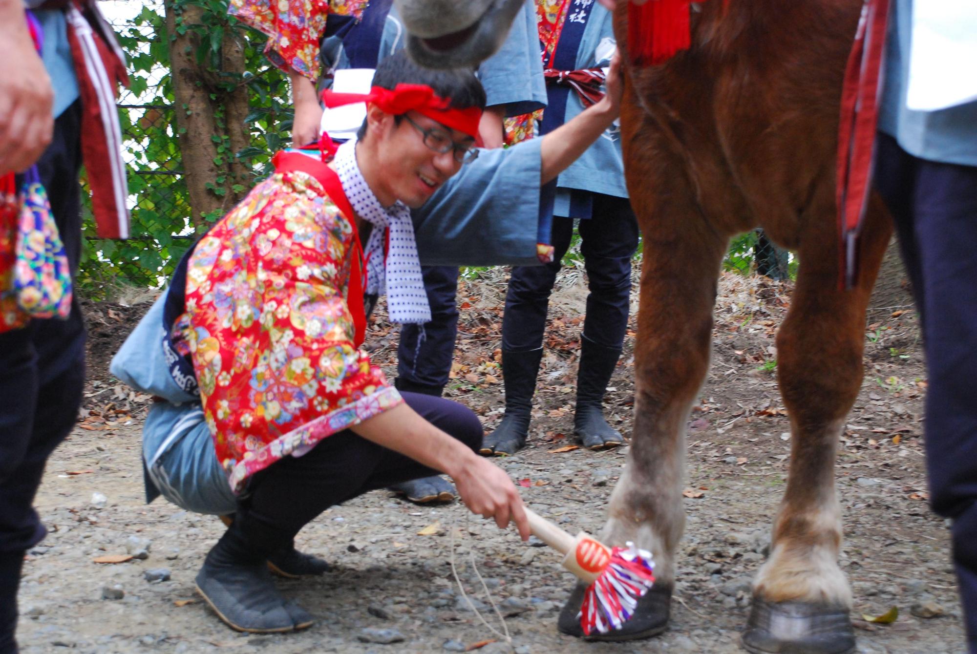 頭に赤い鉢巻を巻き花柄の着物を上にまとった男性が、爪きりの道具を使って馬の蹄を触っている儀式の様子の写真