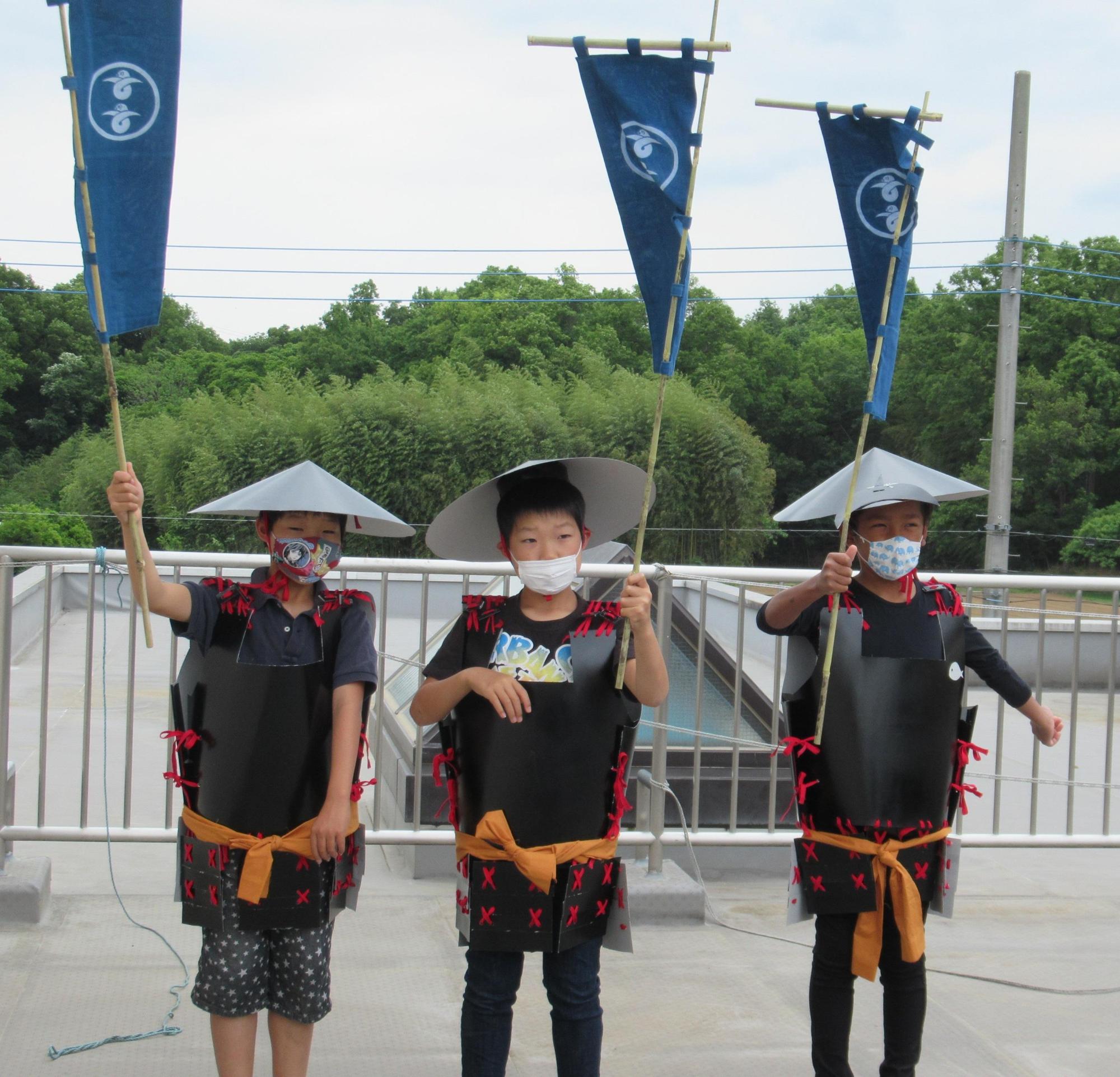 のぼり旗を持ち笠を頭にかぶり、手作りの甲冑を着た小学生3人が並んで立っている写真