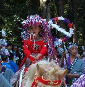 白馬に乗って、頭上にカラフルな色の飾りを乗せ赤色の流鏑馬の衣装を着た男の子の写真
