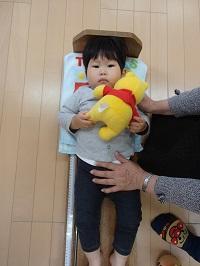プーさんのぬいぐるみを持った男の子が乳児用ベッド型身長計に寝かされ、身長を計測している写真