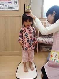 緊張した表情で身長計に立っている女の子と計測してはかりに目を向けている保健師さんの写真