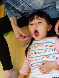 お母さんの膝の上に子どもの頭を乗せている女の子が大きな口を開けて歯ブラシで仕上げ磨きをしてもらっている写真