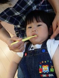 お母さんの膝の上に子どもの頭を乗せて、お母さんが黄色い歯ブラシを持って女の子の仕上げ磨きをしている様子の写真