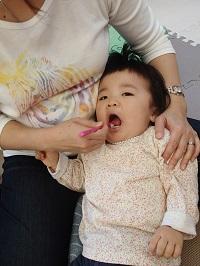 お母さんの膝の上に子どもの頭を乗せて、お母さんがピンク色の歯ブラシを持って女の子の仕上げ磨きをしている様子の写真