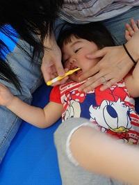 お母さんの両足の間に子どもの頭を置いて、お母さんが黄色の歯ブラシを持って女の子の仕上げ磨きをしている様子の写真