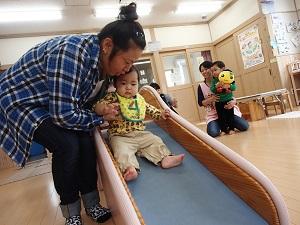 お母さんが赤ちゃんの両脇を抱えながら、滑り台を滑らせて遊んでいる写真
