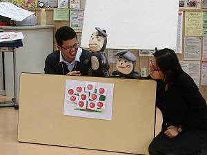 机の台を横に倒してパペット人形の舞台にしており、リンゴのカードが沢山貼ってある木の上で男性と女性がネズミのぱべっと人形を操っている写真