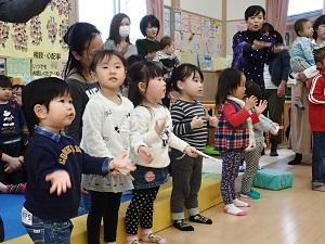 参加者の子ども達が横一列に並んで、両手を広げた同じ動きをして前方を見ており、その後ろで保護者の方々が見守っている写真