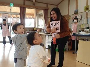 アルファベットが書かれたドキンちゃんのペープサートを持っているサラ先生と向かい合って立っている2人の男の子の写真