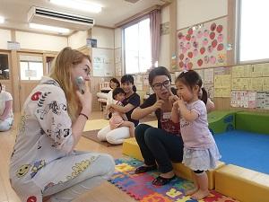 人差し指をほほに当てて立っている女の子と向かい合っているフリーダ先生、女の子の横に座っている母親の写真