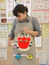 アンパンマンの顔を付けたエプロンをしている歯科衛生士さんが、歯ブラシを右手に持ってアンパンマンの歯磨きをしているエプロンシアターの様子の写真