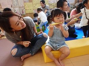ブロックに座った女の子が黄色い歯ブラシを持って前歯を磨いている様子をお母さんが笑顔で見守っている写真
