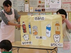 クリーム色の布に貼られた虫歯になりやすい飲み物（コーラ、100パーセントみかんジュースなど）と虫歯になりにくい飲み物（牛乳、麦茶）のイラストを歯科衛生士さんが説明をしている写真