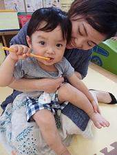 お母さんの膝の上に座っている小さな子供がオレンジ色の歯ブラシを持って歯磨きをしている様子をお母さんが笑顔で見守っている写真