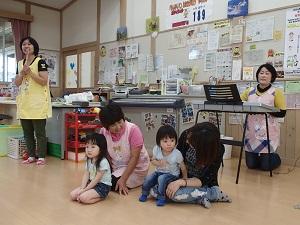 左側に黄色のエプロンを着用した女性スタッフが立ち、右側のエレクトーンの後ろに膝まづく女性スタッフ、前にピンク色のエプロンを着用した女性スタッフの前に女の子が正座し、右隣に膝抱っこしている子供の顔を覗いて見ている写真