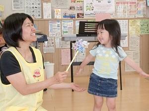 座っている黄色のエプロンを着用した女性スタッフが、右手で棒の先にアジサイやカタツムリが描かれてたイラストに6月と書かれたものを、右側の笑顔の女の子に向けている写真
