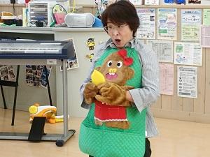赤色のスカートに頭にリボンをつけた大きなサルが緑色のエプロン全体に縫い付けられ、女性が手の部分に両手を入れて読み聞かせを行っている写真