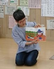床に正座した、赤色の縁の眼鏡をかけた女性が上下に絵本を開き、読み聞かせを行っている写真
