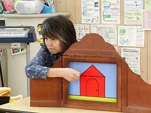 女性が、赤色の三角屋根の家の場面が表示された、木製の枠の紙芝居を行っている様子の写真