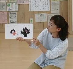 床に正座した、グレー色の服を着た女性が絵本を開いて持ち、読み聞かせを行っている写真