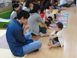 子どもの遊びスペースで参加した親子が座り、手前の青色のシャツを着た母親と男の子が向かい合って座り、母親が両手を口に当てているのを男の子が見ている写真