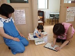 お母さんの横でメモをしている保健師さんの隣でで体重計にのり座っている赤ちゃんの写真