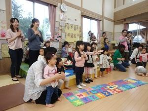 横に並んで手遊びをしている子供たちとその後ろに立っている母親の写真