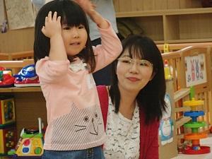 両手を頭の上に置いて立っている女の子と横に座って見ている母親の写真