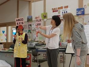 アルファベットが書かれたペープサートを持っている2人の保育士とその横に立っているフリーダ先生の写真