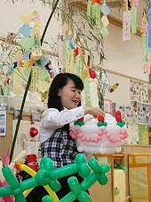 大きな笹の木に短冊が飾られている前で、女性が風船で作った大きなデコレーションケーキを両手で持っている写真