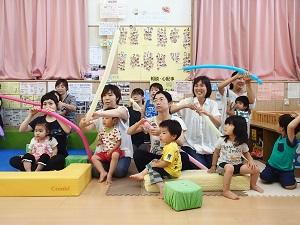 子どもの遊びスペースに参加した親子が座り、カラフルな色の細長く膨らませた風船を母親たちがそれぞれ持っている写真