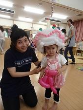 左に膝まづいて座る母親の右側で、水玉柄のピンク色のワンピースを着た女の子が両手でピンク色のバルーンアートを持ち、頭に白色の大きな帽子をバルーンアートで表現したものを頭に被っている写真