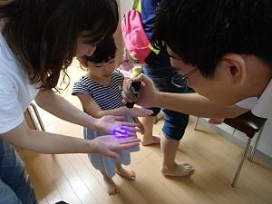 先生がブラックライトで母親と子供の手を照らしてちゃんと手が洗えたか確認している様子の写真