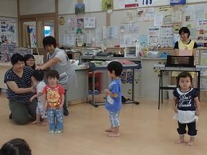 右側にエレクトーンの後ろに立つ黄色のエプロンを着用した笑顔の女性スタッフと、前に3名の子どもが間隔をあけて立ち、左側に母親の前に立つ男の子が後ろを向いてエプロンを着用した女性スタッフに頭を撫でられている写真