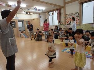 子供の遊びスペースに、参加した親子が座ったり立ったりして、左側に立つエプロンを着用した女性スタッフが右手を上げてグーをしているのを、手前に立つ女の子がカメラに向かってグーのポーズをとっている写真