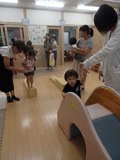 母親に付き添われながら木製の平均台を降りようとしている女の子、手前の小さなすべり台に母親の手を借りて登ろうとしている男の子の写真