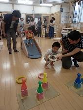 父親が見守るなか、すべり台から滑ろうとしているアンパンマンのシャツを着た男の子、手前のカラフルな色のボウリングのピンに向かって、母親から渡された輪投げの輪を受け取ろうとしている男の子の写真
