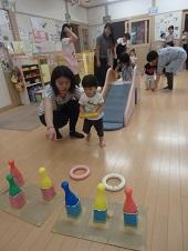 母親に付き添われながら木製の平均台から降りた子供、手前の小さなすべり台を母親に見守られながら登っている女の子、手前のカラフルなボウリングのピンに向かって、母親から教わりながら黄色の輪を投げようとしている男の子の写真
