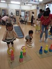カラフルな色のボウリングのピンに向かって黄色の輪を投げている女の子、右側の母親が見守るなか白色の輪を拾っている男の子の写真