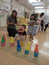床に膝をついて座っている母親に見守られながら、カラフルな色のボウリングのピンの手前でピンク色の輪の穴からピンを見ている男の子、隣で白色の輪を持っている女の子の写真